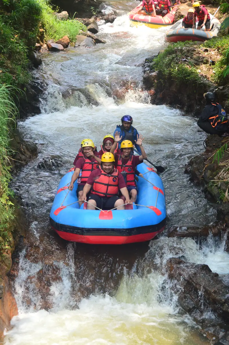 Rombongan PT Antarmitra Sembada memasuki jeram pertama Sungai Palayangan; guide FAST Rafting berdiri mengendalikan perahu dari belakang sementara peserta berseragam merah maroon duduk berpegangan sambil melambaikan tangan.
